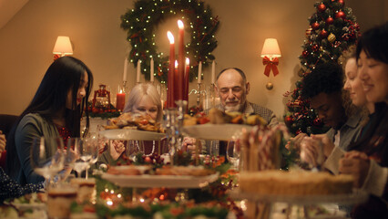 Happy large family praying before Christmas, then starting to eat and celebrating. Served holiday table with delicious meal and candles. Warm atmosphere of family Christmas dinner.