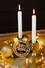Candles in bronze candlesticks, bright balls decoration for Christmas tree and wooden plate with Merry Christmas lettering on a golden stand. Black wooden background. Candle lights and led flashlights