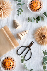 Fototapeta na wymiar Fragrant pomander balls handmade from tangerines with cloves. Handmade paper stars from brown baking paper. Flat lay on off white textile tablecloth with eucalyptus. Wood Christmas tree toys, trinkets