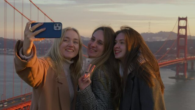 Female friendship, travel together to iconic tourist destinations concept. Portrait of caucasian women taking photos on golden hour lighting by the iconic, suspension bridge. High quality 4k footage