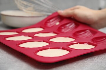 Woman holding mold for madeleine cookies with batter at grey table, closeup