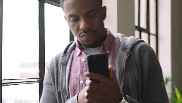 Black man, phone and coffee drink by window in office for digital networking, social media or reading morning news online. African man, smartphone and wifi network communication or typing email