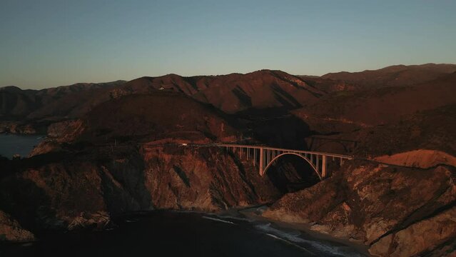 Scenic, winding road by the edge of a ocean cliff as seen from above. Aerial view of the Bixby Creek Bridge lit by the golden hour lighting. High quality 4k footage