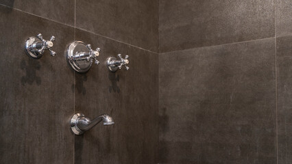 Corner fragment of a shower stall. On the black tiled wall there are metal valves for hot and cold water, a faucet.