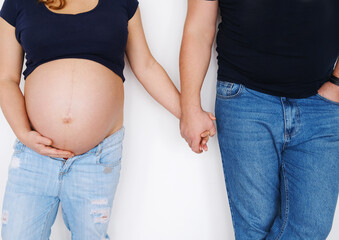 A couple, a man and a pregnant woman stand near a white wall and hold hands. The concept of support and love in the family.