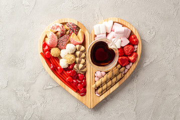 Valentine's Day celebration concept. Top view photo of wooden heart shaped serving tray with sweets chocolate jelly candies cookies and glass cup of drinking on concrete texture background