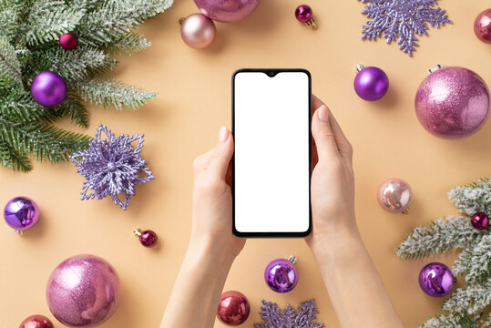 Christmas Day concept. First person top view photo of girl's hands holding smartphone over pink violet baubles flower snowflake ornaments fir branches on isolated light beige background with copyspace