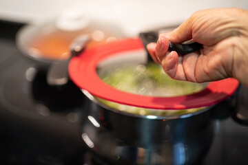 Female hand open pan lid on electric hob with boiling water or green soup and scenic vapor steam...