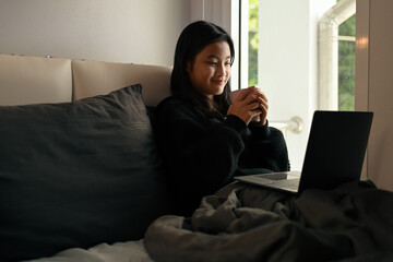 Smiling asian girl drinking hot chocolate and using laptop on bed, relaxing on weekend morning