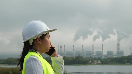 Female chief engineer in green vest and helmet talking on the phone outside against the background of coal power plant station and steam in the morning mist.