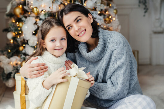 Cheerful brunette female leans at her daugter, embraces her, presents gift box, being in living room near decorated New Year tree. Glad family: mother and daughter in warm sweaters celebrate Christmas