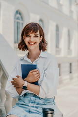 A woman in a white shirt and a blue denim skirt sits on a parapet and cuddles a blue notepad against a building. Summer walks in the city, travel.