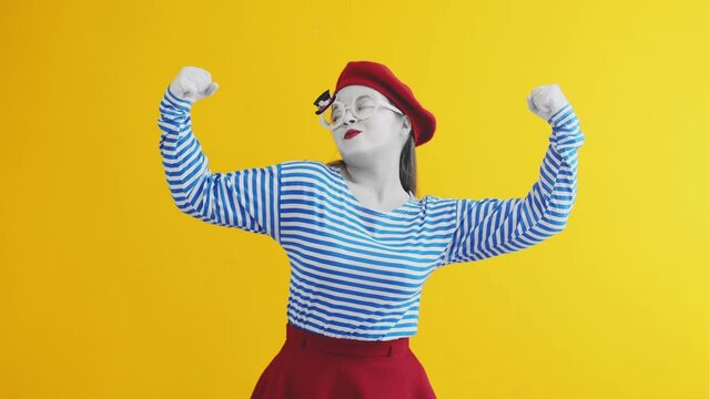 Young mime woman self-confidently demonstrating her strength by showing her biceps. Mime playing funny performance on yellow background