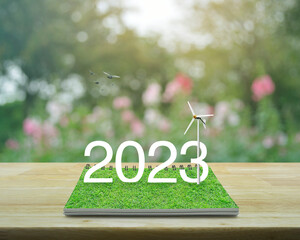 2023 white text with wind turbine on green grass on open book on wooden table over blur pink flower...