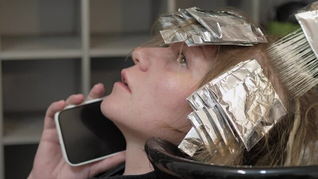 Busy woman talking on the phone while dyeing her hair by a professional master in a beauty salon with a special sheet of foil