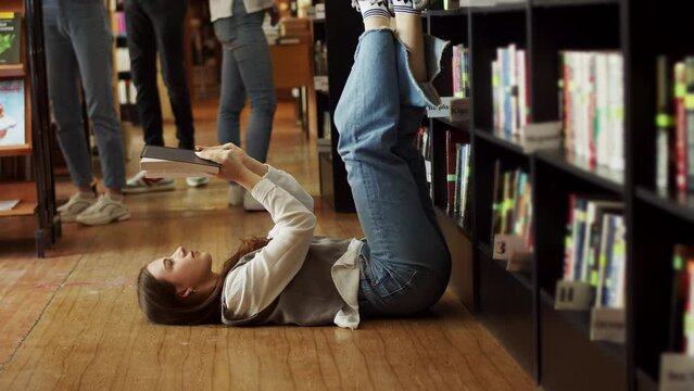A student girl lies on the floor in the university library, legs up. Caucasian woman holding a book in her hands. In the background are bookshelves and a group of young people.