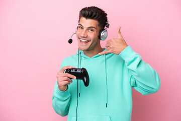 Young handsome caucasian man playing with a video game controller isolated on pink background making phone gesture. Call me back sign