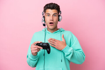 Young handsome caucasian man playing with a video game controller isolated on pink background surprised and shocked while looking right