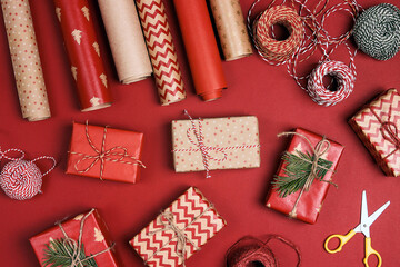 Christmas preparations with wrapped presents, decorative ropes, paper roll and scissors on red background.