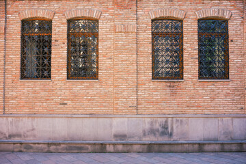 Facade of a brown brick wall with four windows and sidewalk in the foreground. Front view without...