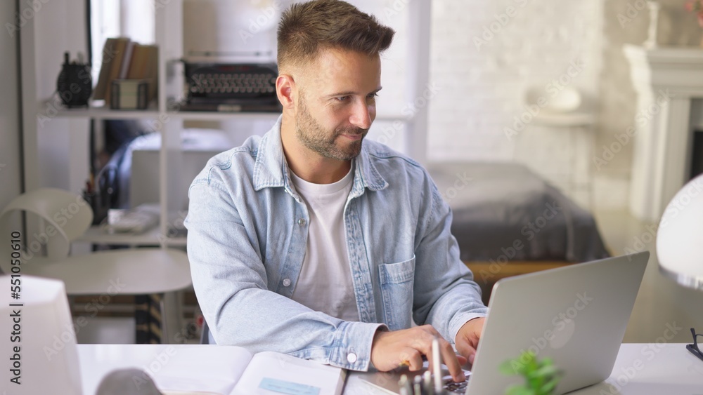 Wall mural Happy businessman in casual using laptop in home office, Young adult man sitting at desk in study room, working online with computer, browsing the Internet, smiling.