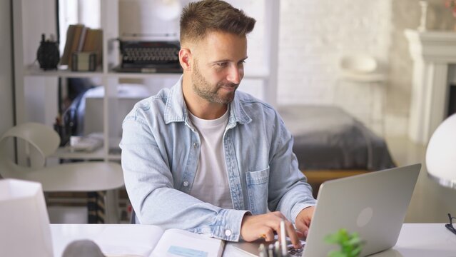 Happy businessman in casual using laptop in home office, Young adult man sitting at desk in study room, working online with computer, browsing the Internet, smiling.