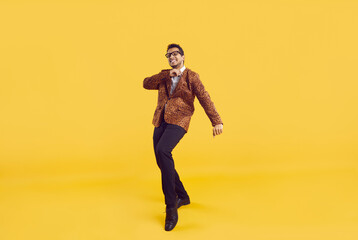 Fototapeta na wymiar Funny young black guy in a party outfit dancing alone on a yellow colour background. Goofy positive African American man in a leopard jacket and glasses enjoying music and having fun in the studio