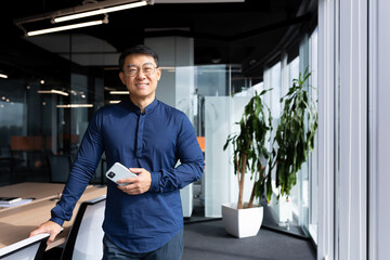 Portrait of successful Asian businessman, man in glasses and shirt smiling and looking at camera, successful investor holding phone standing near window.