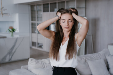 Fototapeta premium Stunning caucasian model relaxing at home sits on cozy couch eyes closed touches her perfect long hair satisfied by her hair care products and shampoo. Attractive Italian young woman feels headache.
