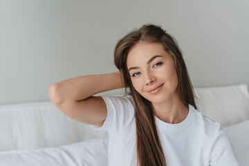 Adorable caucasian female model looks at camera with cute smile relaxing at home sits on bed. Close up portrait of gorgeous Italian girl with perfect skin and hair. Healthy lifestyle and beauty model.