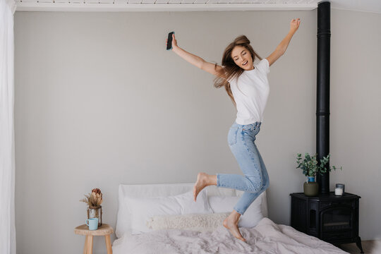 Jumping Hi Young Adult Italian Woman In White T-shirt And Blue Denim Jeans Holds Phone Happy Jumps On Bed At Home. Mockup With Copy Space With Celebrating Caucasian Girl. Happiness And Youth.