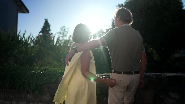 Back Of Joyful Senior Couple Embrace Standing Outside Looking At Sun. Tender Moment Between Older Married Man And Woman With Arm Around Standing At Park