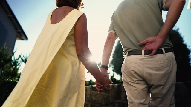 Back of two older people holding hands together standing outdoors in sunlight. Senior couple with hand held together. Resilience love and support concept