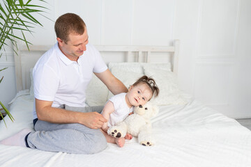 father plays with baby daughter teddy bear on white bed at home, happy family, father's day