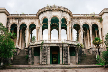 Fototapeta premium Facade of a decaying palace or hotel with tall marble columns and arches. The abandoned building is surrounded by bushes against a gray sky