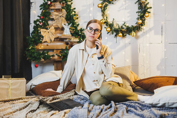 Cute young woman talking on phone to prepare for Christmas with laptop and technology while sitting on bed near Christmas tree at home on holiday.
