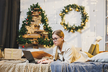 Young beautiful woman in cozy home clothes using laptop on bed in room with Christmas tree. Portrait of carefree happy girl student at home in Christmas