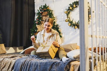 Happy pretty woman with fair hair in cozy clothes with a New Year's gift in her hands is sitting on the bed near the Christmas tree. New Year's atmosphere at home.