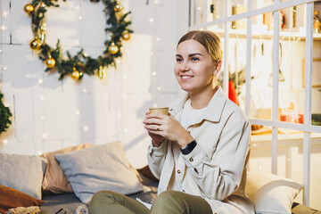 Portrait of a smiling woman with fragrant coffee in her hands against the background of New Year's decorations. Festive Christmas coffee at home on the bed, New Year holidays