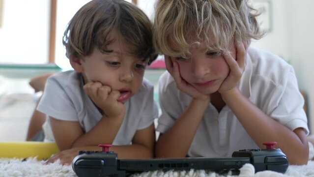 Two small boy with hand in chin watching media online with tablet device. Children lying on floor at living room home staring at technological screen