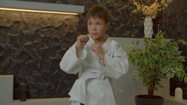 Concentrated determined cute multiracial karate kid in kimono practicing karate defensive skills, performing and improving elbow block techniques while doing indoor sport training.