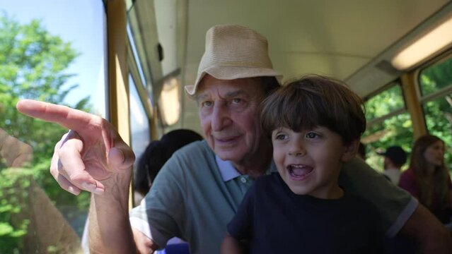 Happy grandfather traveling by train with grandchild together. Senior man pointing at landscape inside railroad wagon in motion. Family on vacation