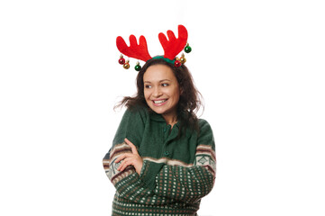 Multi-ethnic joyful attractive woman, wearing deer antler hoop and warm green sweater with Christmas pattern, hugging herself, laughing, looking aside at copy advertising space on white background