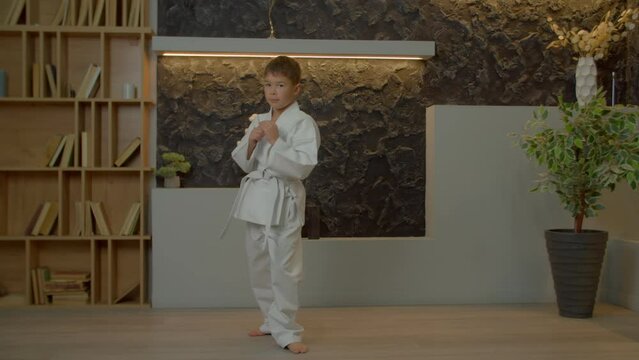 Determined motivated cute multiethnic elementary age karate fighter in white kimono training karate techniques, performing and improving roundhouse kick while practicing in domestic room.