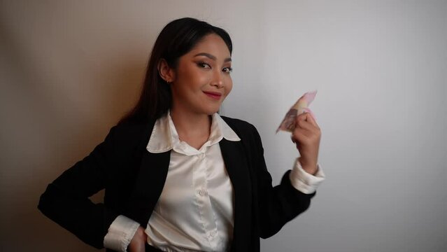 A happy young businesswoman is wearing black suit and holding cash money in Indonesian rupiah isolated by white background