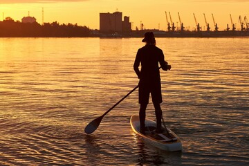 Silhouette of a man with a paddle on a SUP (stand up paddle board) at early sunny morning on river against orange dawn of port cranes