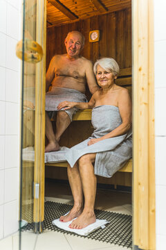 Vertical Indoor Shot Of Married Couple Enjoying Sauna. Caucasian Senior Woman In Gray Towel Touching The Knee Of Her Male Partner Sitting On Higher Bench. High Quality Photo