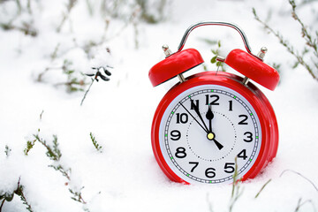 A red retro alarm clock showing 12 o'clock is standing in the snow. New Year, Christmas.