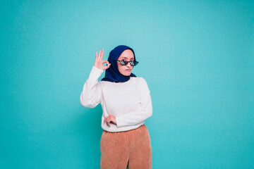 Fototapeta premium Portrait of a smiling Asian Muslim woman wearing a glasses, giving an OK hand gesture isolated over blue background