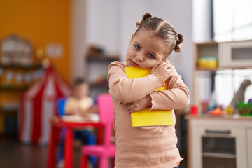 Adorable hispanic girl student hugging book standing at kindergarten